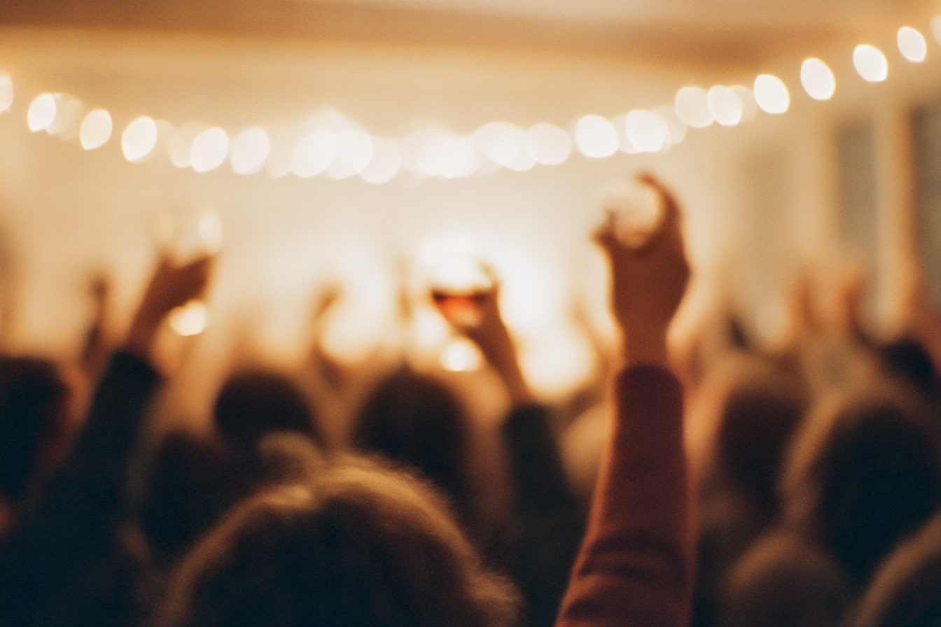 A warm, crowded gathering — string lights, raised glasses, faces turned away